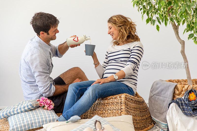 深情的男人倒新鲜牛奶在马克杯为他的愉快怀孕的妻子坐在客厅的Hassock -库存照片